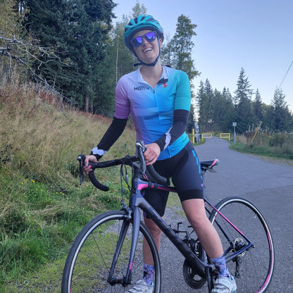Woman smiling while standing astride on a bike, wearing sunglasses, a helmet, and a colorful MōTTIV cycling jersey, and shorts.
